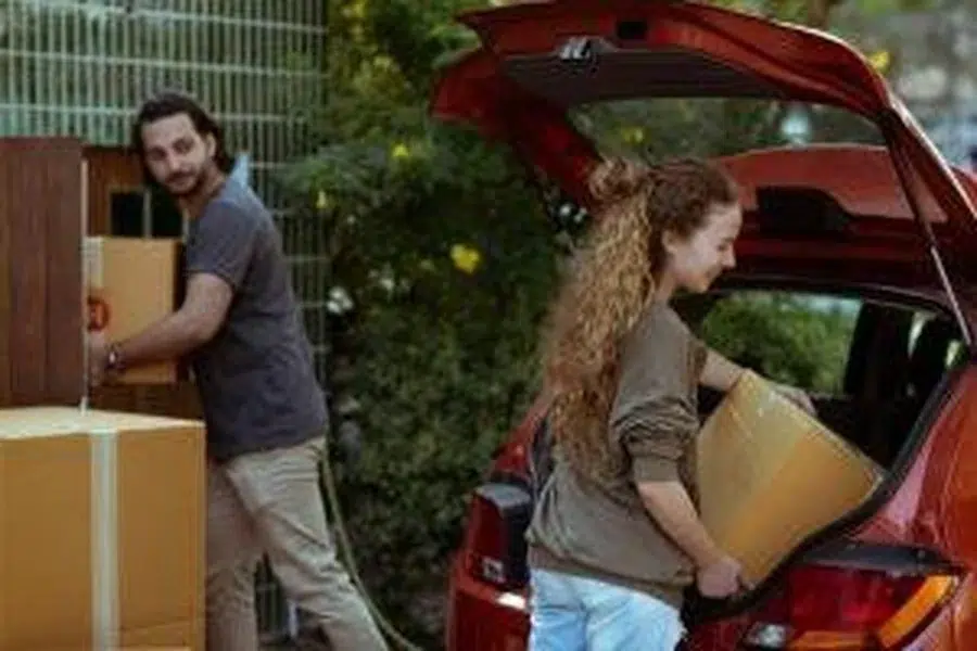 A woman unloads a box from a car while a man carries boxes toward a new home surrounded by greenery.