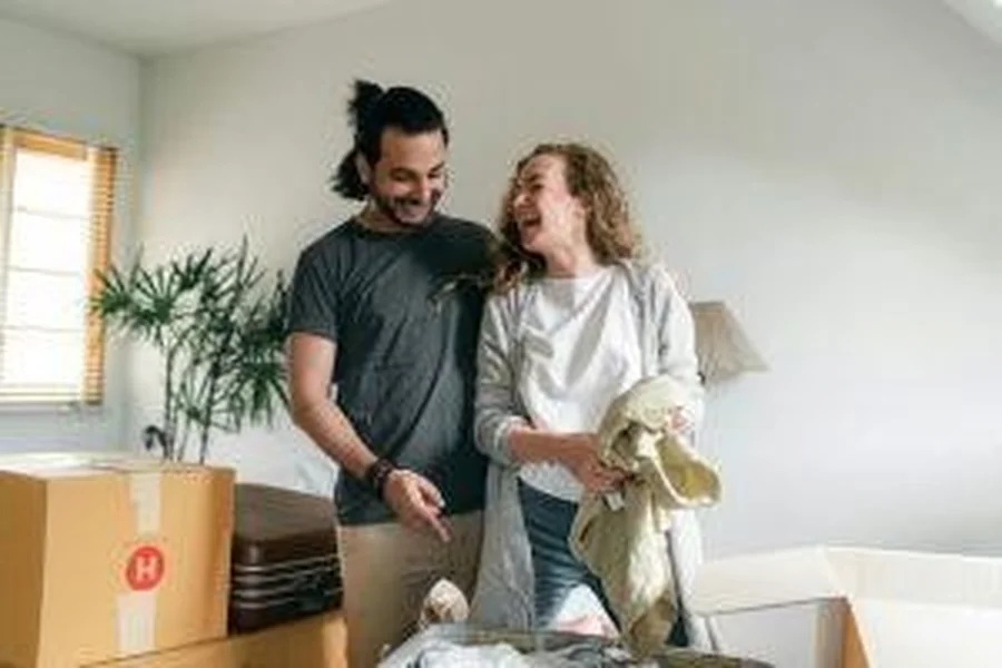 Tag: A smiling couple unpacks clothes from boxes and a suitcase in their new home, sharing a light moment together.